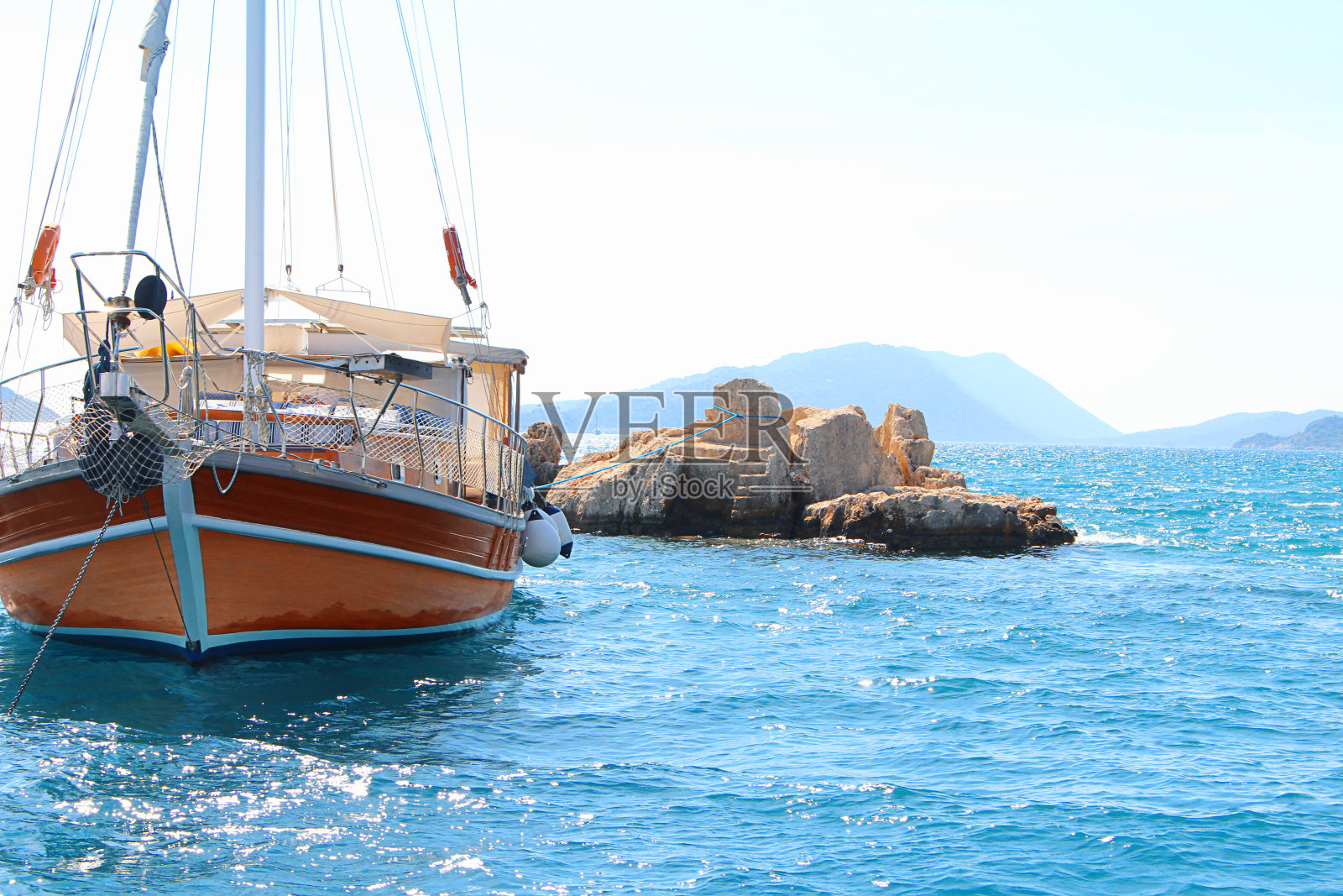 在晴朗的阳光明媚的日子里，折叠帆的白色和棕色单桅帆船停泊在公海的岩石上。船首甲板、桅杆和风帆在平静的海面上照片摄影图片