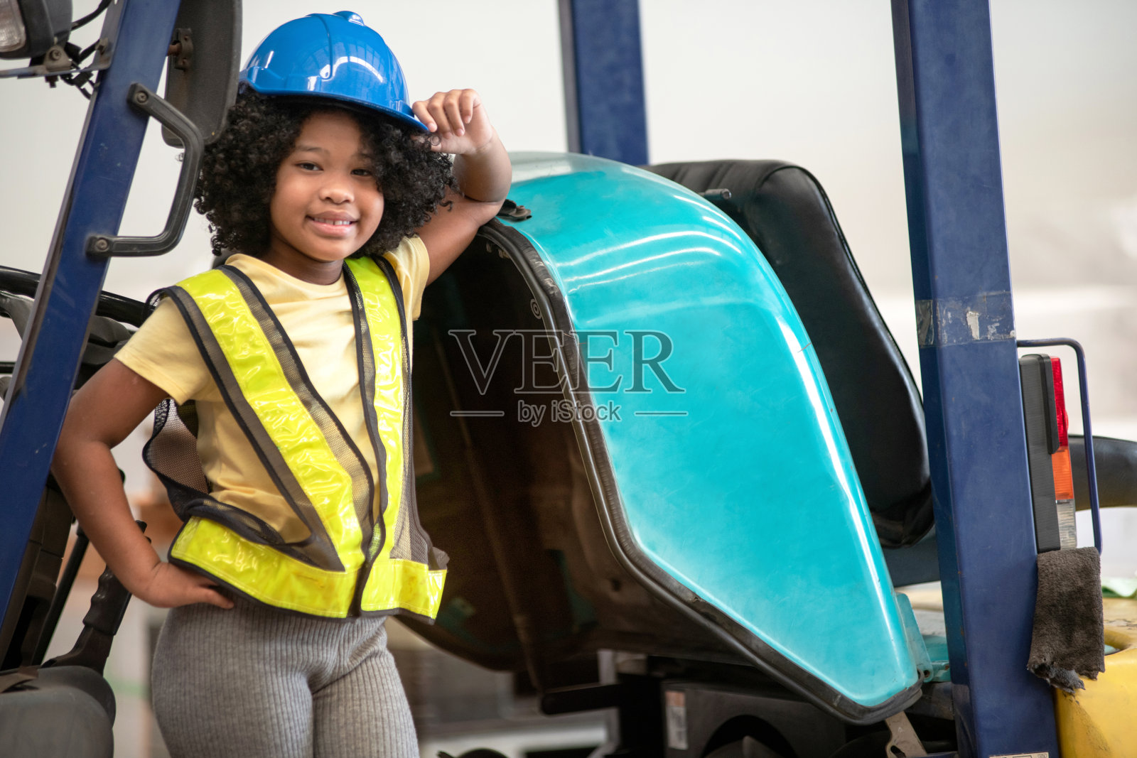 一个非洲小女孩穿着反光背心和安全帽站在仓库的叉车旁边照片摄影图片
