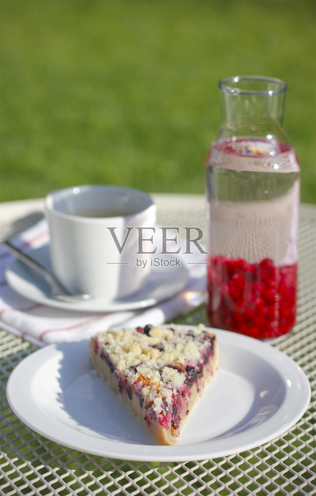 一块自制的蛋糕，在白色的盘子里放着红、黑加仑子和面包屑，一杯黑咖啡，配有茶托和茶匙，加仑子柠檬水和条纹茶巾，放在白色的花园桌子上，背景是新鲜的绿草照片摄影图片