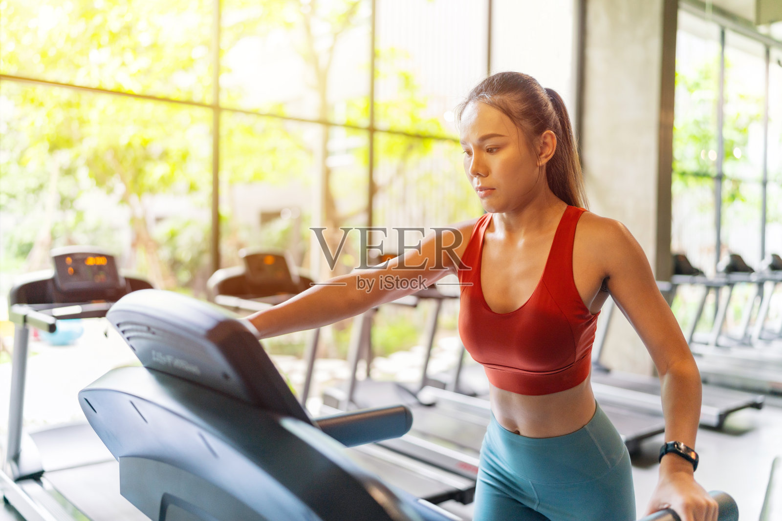 亚洲女性在阳光下跑步，透过窗户在室内健身俱乐部与花园景观照片摄影图片