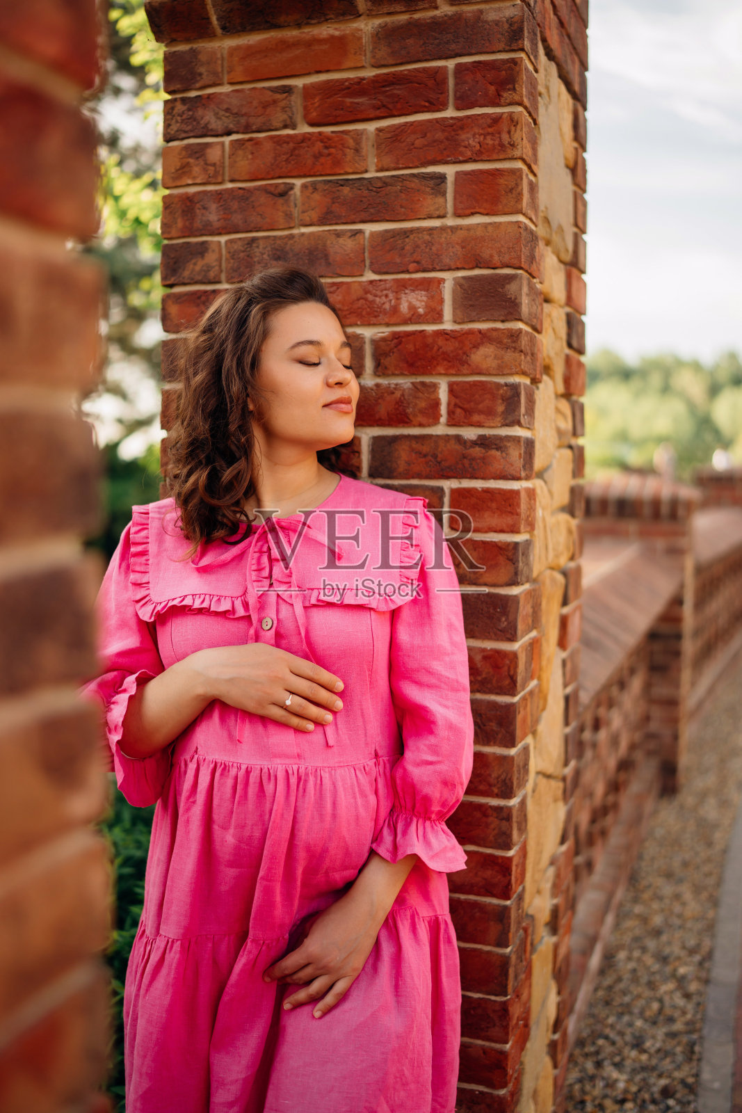 幸福的年轻孕妇穿着粉红色的裙子，抱着她的肚子站在石栅栏旁照片摄影图片