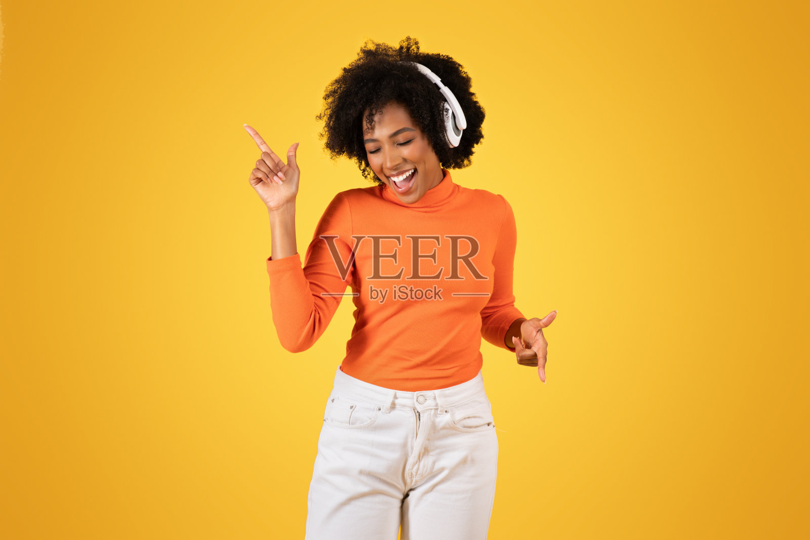 快乐的年轻女子，一头卷曲的非洲式头发，戴着耳机，一边跳舞一边向上指照片摄影图片