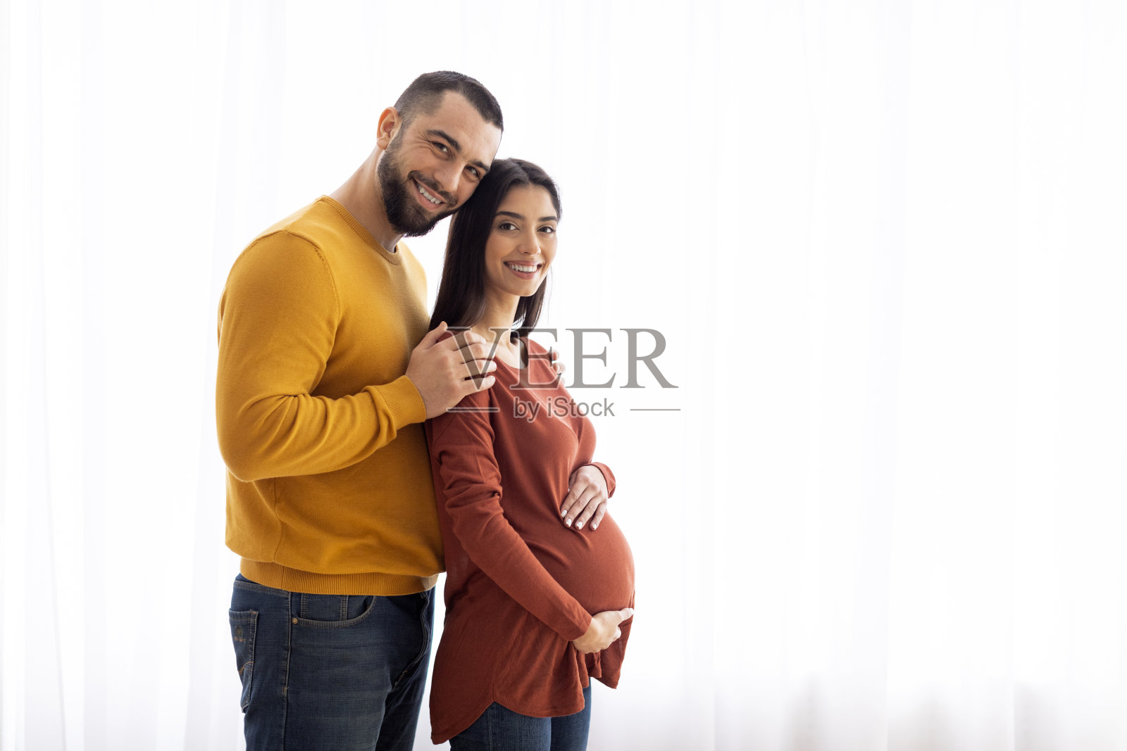 怀孕的白人夫妇站在家里的窗户边拥抱照片摄影图片