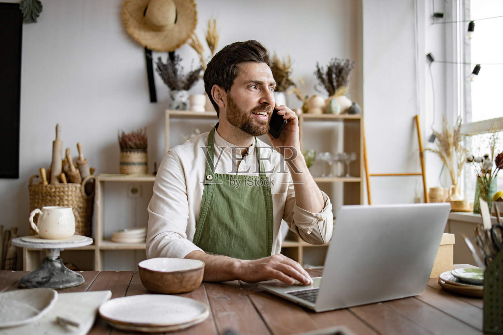 穿着围裙的白人男子坐在陶瓷店的桌子旁，用笔记本电脑工作照片摄影图片