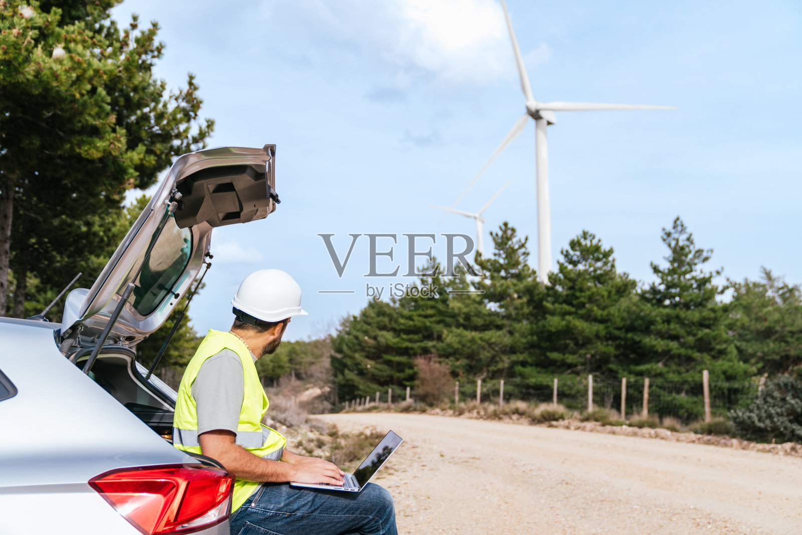 技术人员坐在汽车后备箱上，在土路上靠近风力涡轮机的地方用笔记本电脑工作照片摄影图片