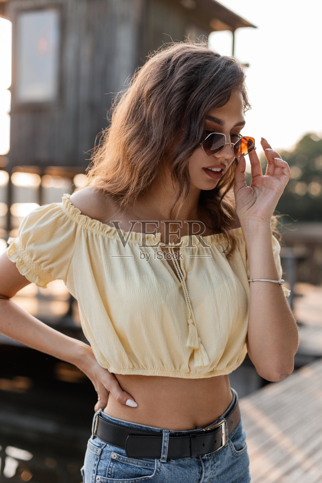 在海滩附近的码头上，时尚漂亮的年轻女性穿着时髦的黄色衬衫和蓝色牛仔裤，戴着时髦的太阳镜。照片摄影图片