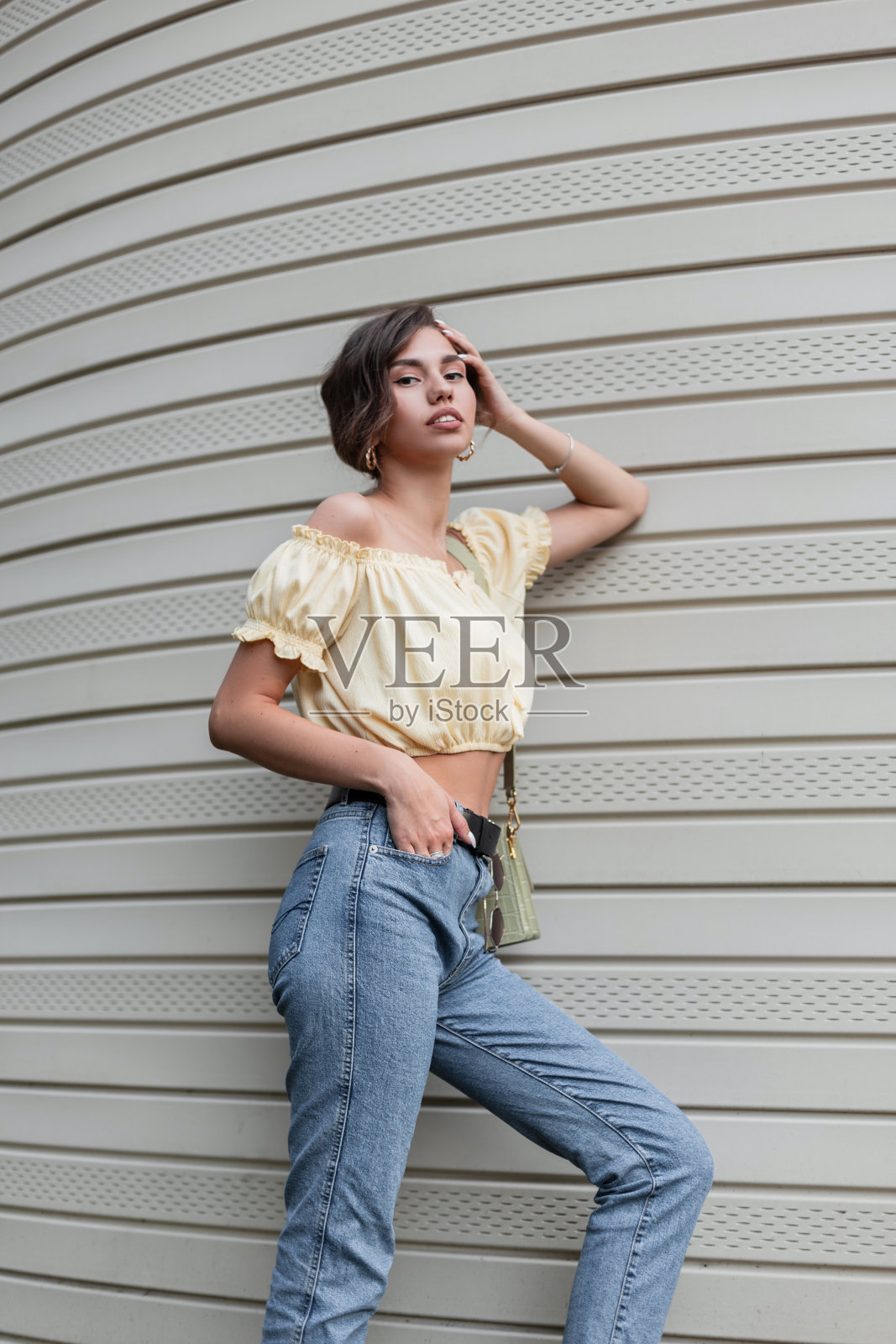 时髦的漂亮女人，穿着夏季的黄色时髦衬衫和复古的蓝色高腰牛仔裤，拿着手提包站在墙边照片摄影图片