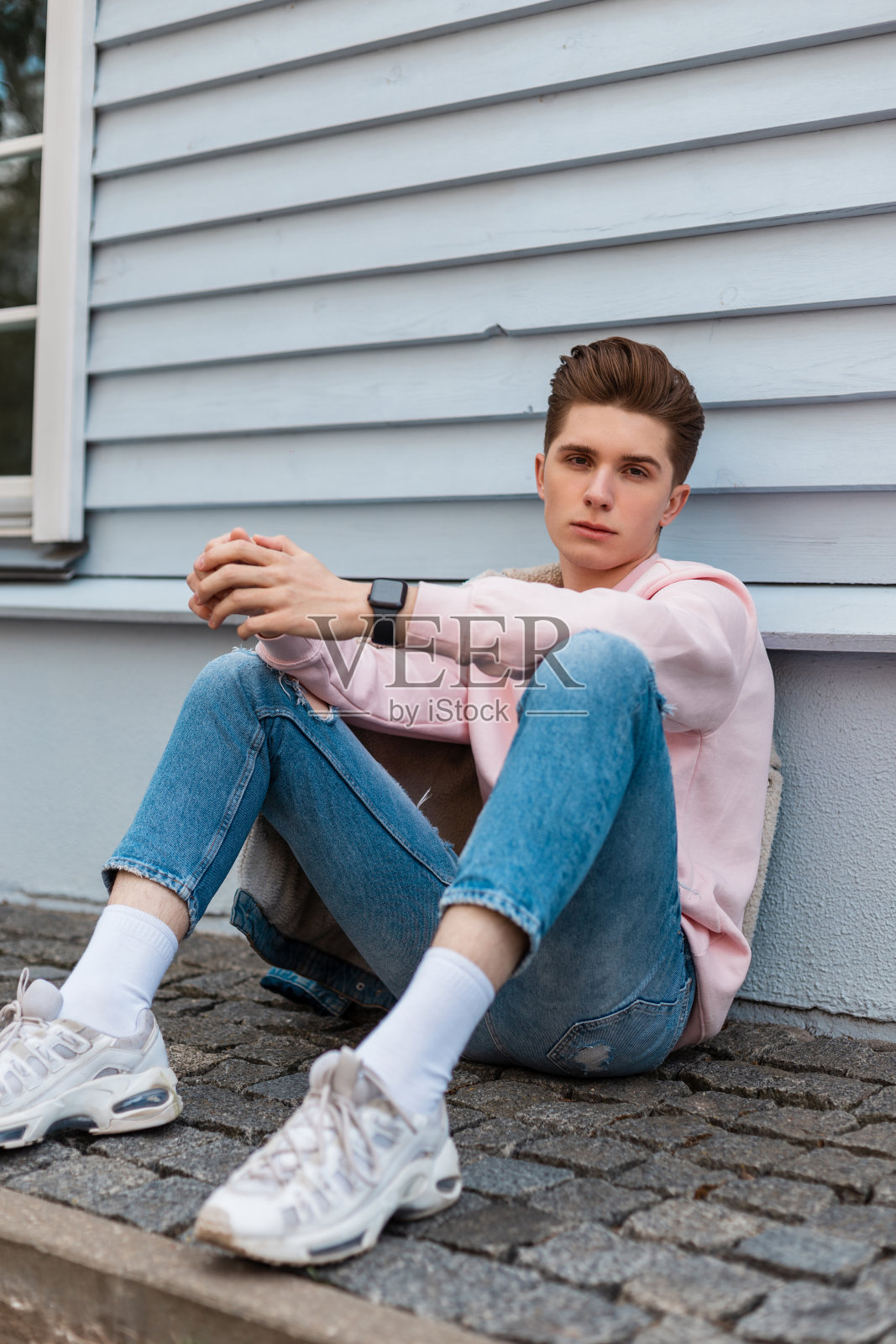 一个时髦的年轻人穿着时髦的粉色运动衫，蓝色时髦的牛仔裤和白色运动鞋，坐在城市街道上一座古老建筑附近的石头瓷砖上。在户外穿着时尚青年服装的帅哥。照片摄影图片