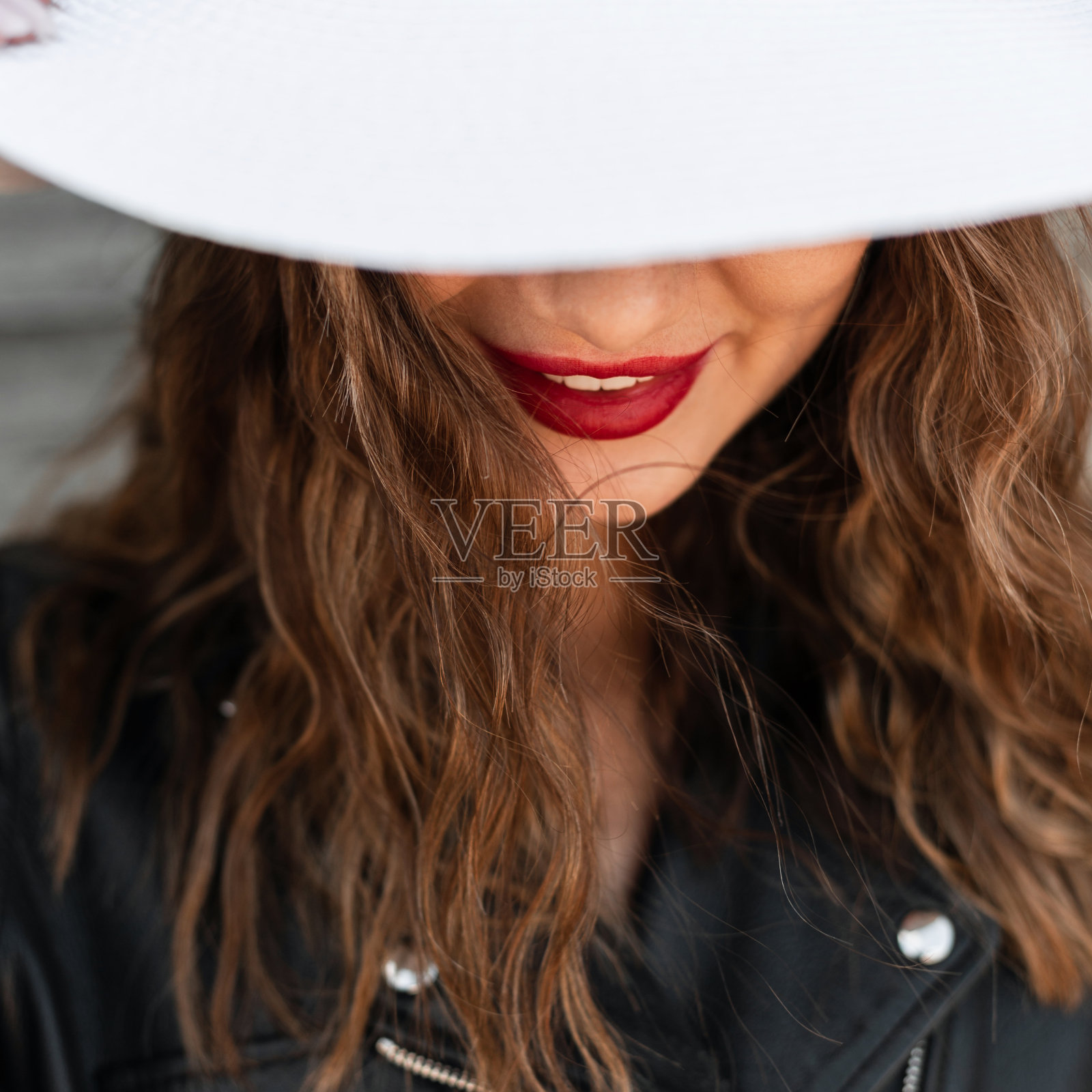 特写肖像快乐的女人，美丽的红唇和积极的微笑，卷曲的棕色头发，黑色夹克，戴着帽子。优雅时尚的白色帽子遮住女性的脸。照片摄影图片