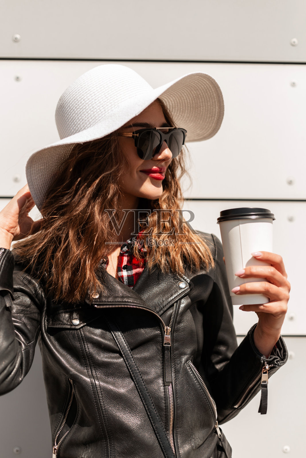 时髦的女孩穿着时髦的衣服，戴着太阳镜，戴着时髦的帽子，在白墙附近的街道上喝咖啡照片摄影图片
