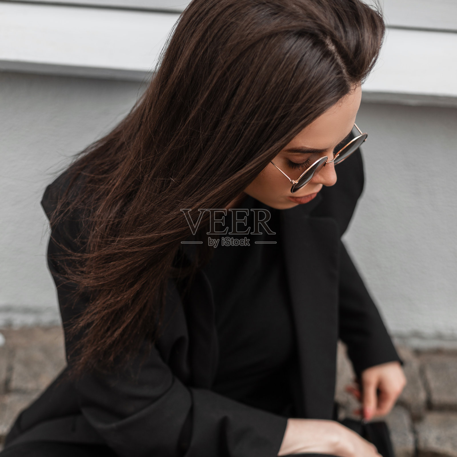 漂亮时髦的年轻女子穿着时髦的黑色衣服，戴着时髦的太阳镜，坐在石头路上，向下看外面。肖像迷人的现代女孩。女性的时尚服装。休闲风格。照片摄影图片