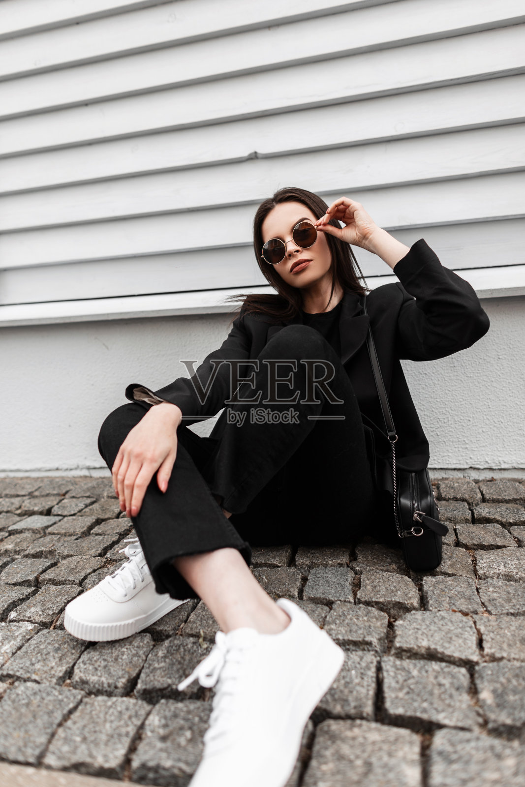 漂亮的年轻女子穿着运动上衣，t恤，牛仔裤，运动鞋，手拿手提包，戴着太阳镜，坐在街上一座古老的木制建筑旁。美丽的女孩穿着时尚的休闲青年装在户外摆姿势。照片摄影图片