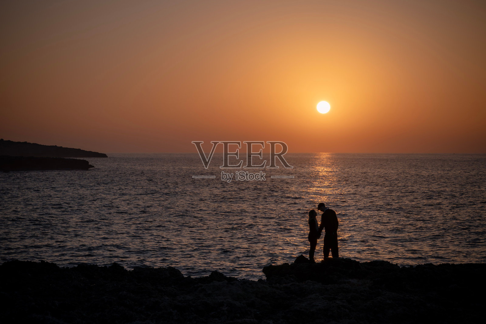 两个人站在海边的岩石上，面对着太阳照片摄影图片