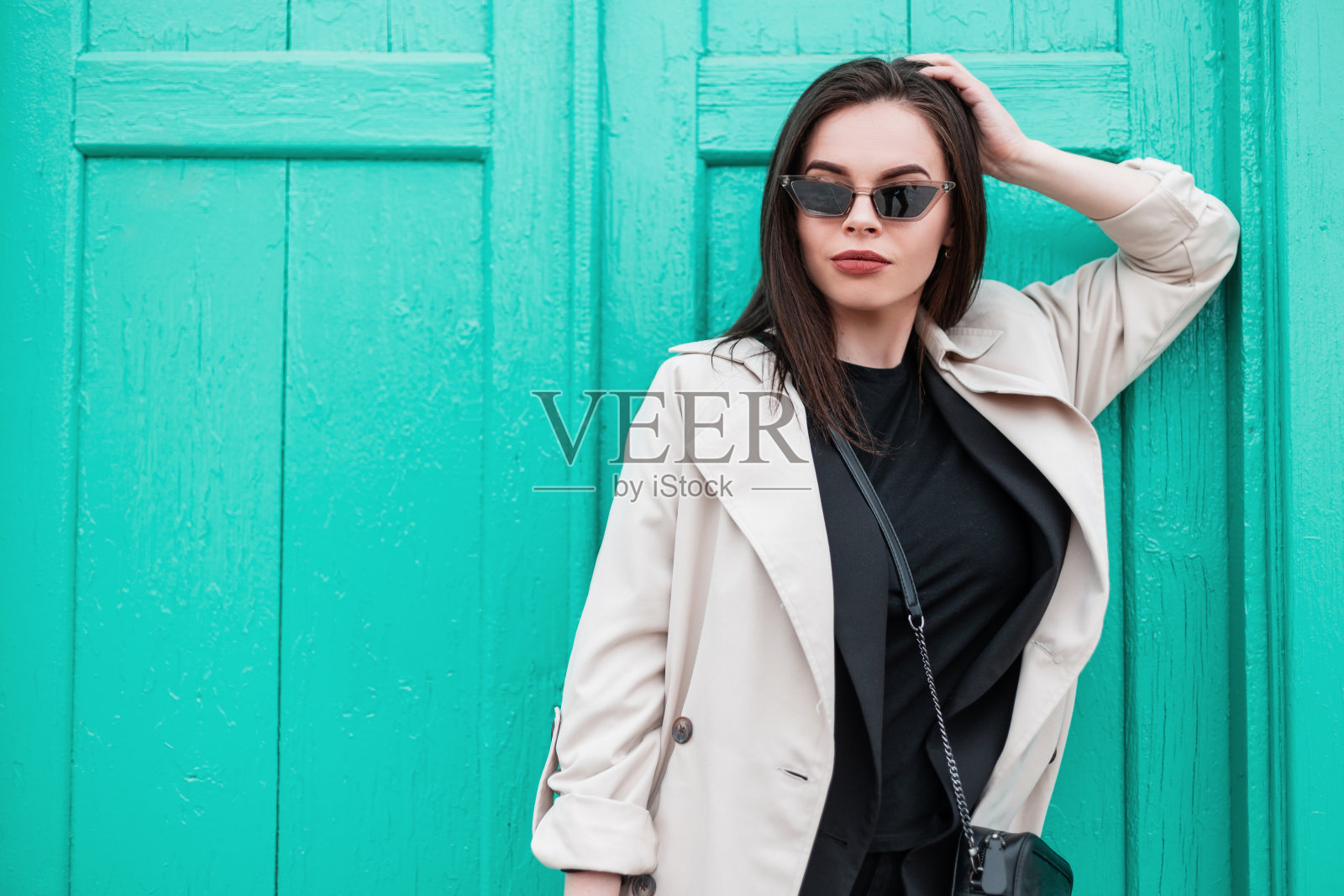 漂亮的年轻女子戴着时尚的复古太阳镜，穿着黑色时髦的t恤，穿着时尚的春季风衣，在街上的复古明亮的绿松石木制门附近休息。都市女孩户外时尚模特。照片摄影图片