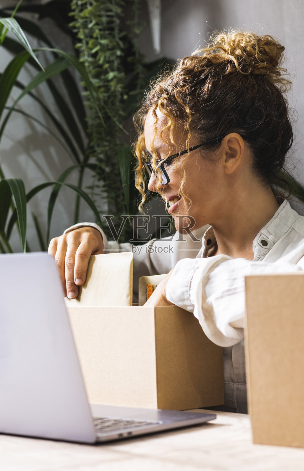 快乐的女人为新成立的家庭公司准备包裹和货物。在办公室工作。办公桌上有包和笔记本电脑的员工。现代忙碌的女士带着微笑工作照片摄影图片