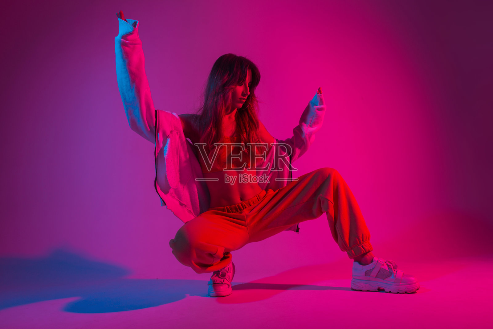 迷人的年轻女舞者穿着时尚的青年服装，穿着白色运动鞋，坐在明亮的粉红色灯光的房间里。现代女孩模特在工作室摆出霓虹灯惊人的彩色红色。照片摄影图片