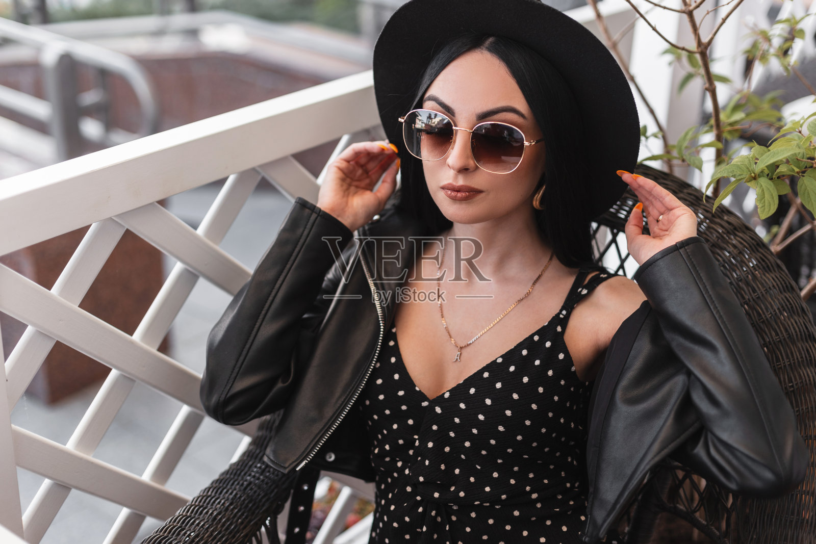 都市华丽的女人戴着时尚的太阳镜，穿着漂亮的黑色连衣裙和皮夹克，拉着复古的帽子。可爱的时尚模特女孩穿着黑色时髦的衣服，坐在咖啡馆的复古椅子上休息。照片摄影图片