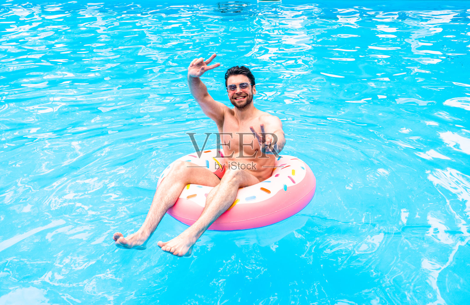 微笑的年轻人在镜头前摆姿势，展示胜利的手势，在游泳池里休息，带着充气戒指。夏天，假期，生活方式的概念照片摄影图片
