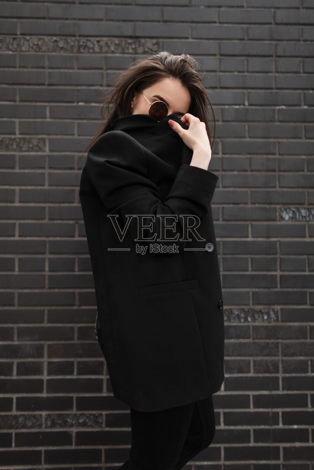 美国时髦的年轻女子，戴着复古风格的时尚圆形太阳镜，盖着时髦的夹克，站在城市的黑砖建筑旁。美丽的都市女孩模特在户外休闲青年服装。照片摄影图片