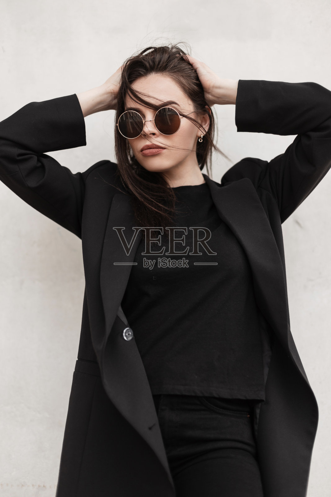 时尚的年轻女子戴着凉爽的圆形太阳镜，穿着时尚的黑色休闲服，在现代的白色墙壁旁拉直发型。美国时尚女孩在街头穿着休闲装。青春的风格。照片摄影图片