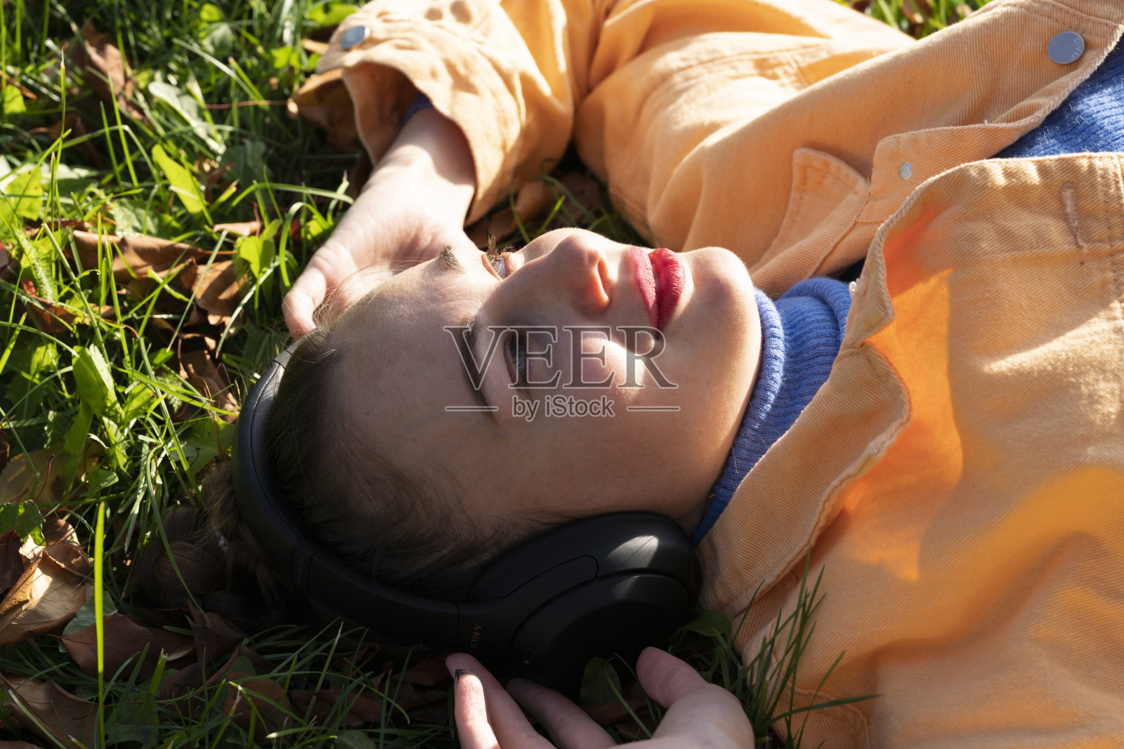 一位年轻女子躺在公园的绿草地上，戴着耳机欣赏音乐。照片摄影图片