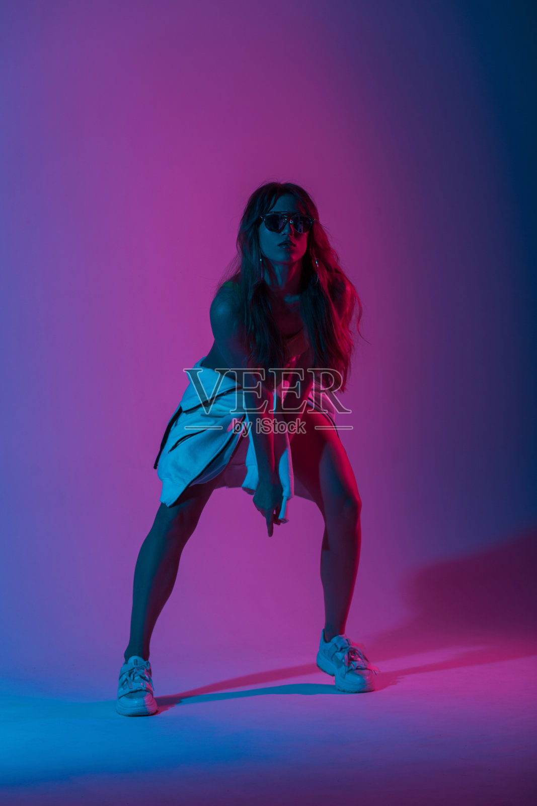 年轻女舞者戴着太阳镜，穿着青年时尚的连帽衫，穿着时尚的运动鞋，在明亮的蓝色霓虹灯下跳舞。女孩在舞蹈棚里欣赏着五彩缤纷的舞蹈照片摄影图片