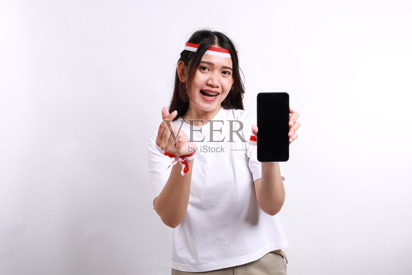 一个快乐的亚洲女人戴着红白发带，展示着她的手机，用手指做着爱的手势，表现出温柔的感觉，被白色的背景隔离。印尼独立日照片摄影图片