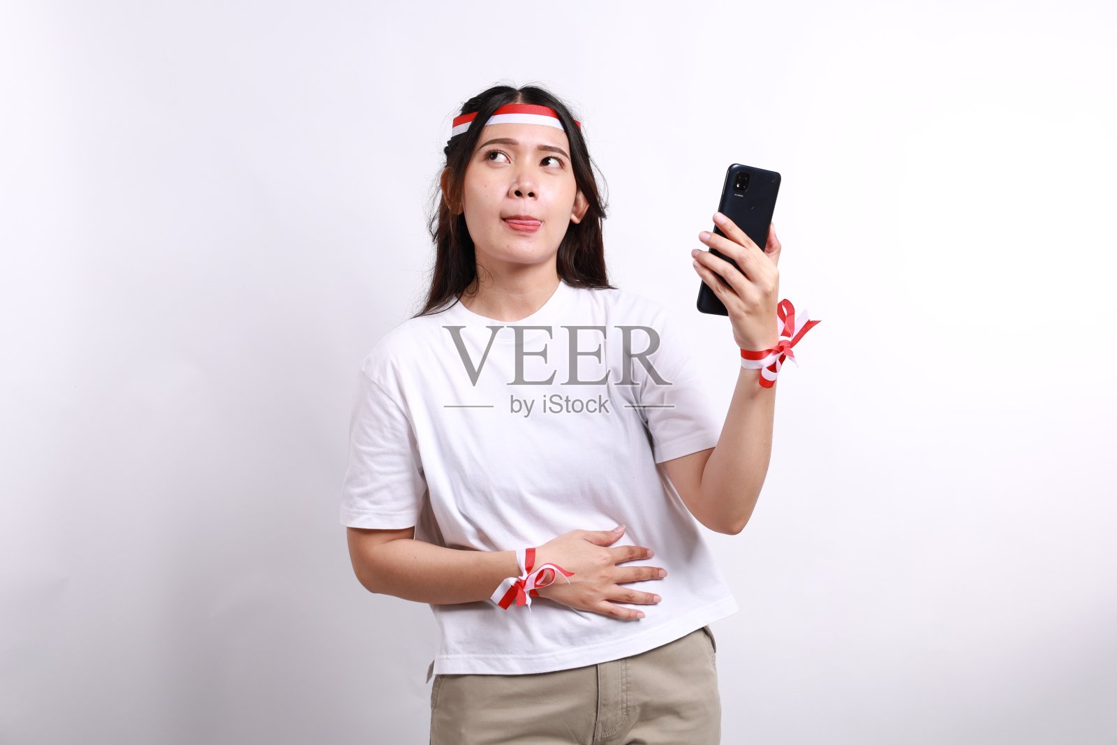 一个体贴的亚洲女人，戴着红白发带，拿着手机和肚子，想着要吃什么或点什么，被白色的背景隔离。印尼独立日照片摄影图片