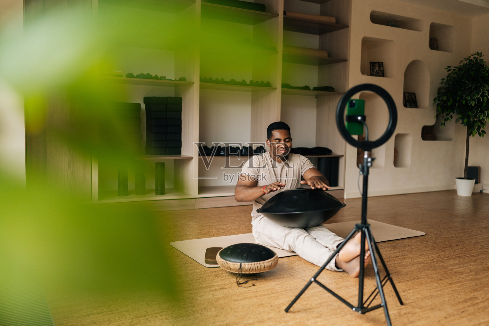 快乐微笑的黑人男博主坐在地板上练习本土音乐传统的鼓乐器，手机前有戒指灯。照片摄影图片