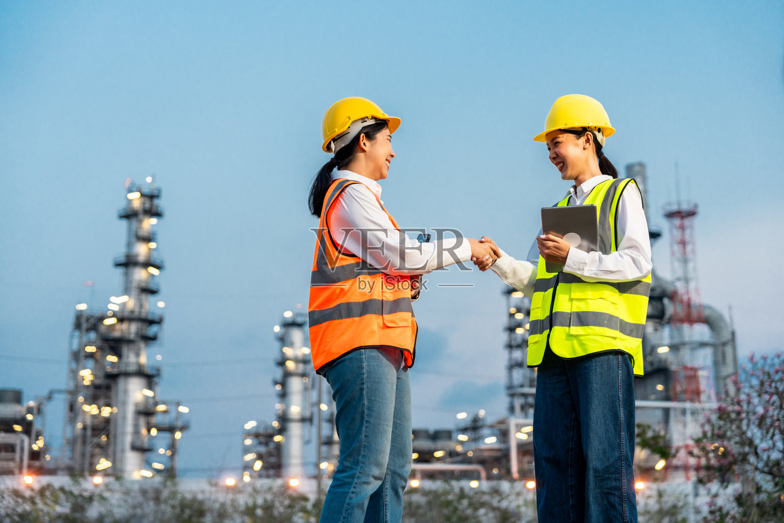 两个戴着白色安全帽的亚洲女工程师站在炼油厂前。工业园区天然气石化。工厂储油罐和管道。工人们在炼油厂施工照片摄影图片