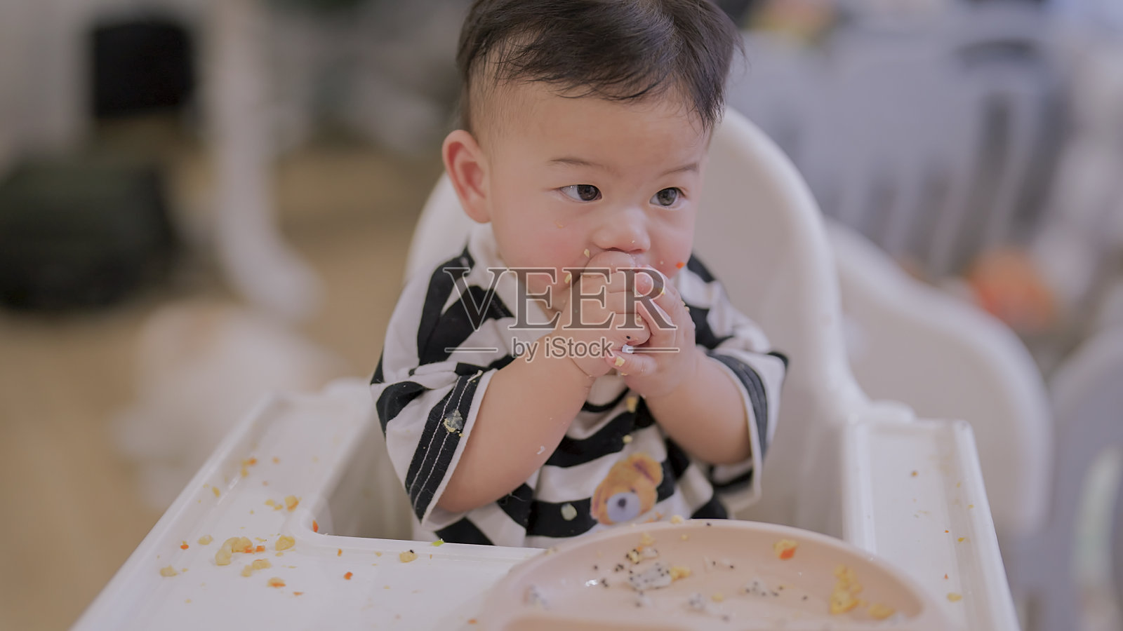 可爱的亚洲男婴坐在高脚椅上自己吃盘子里的食物，母亲的手在家里用勺子喂她的男婴，婴儿里程碑，成长和发展的概念照片摄影图片