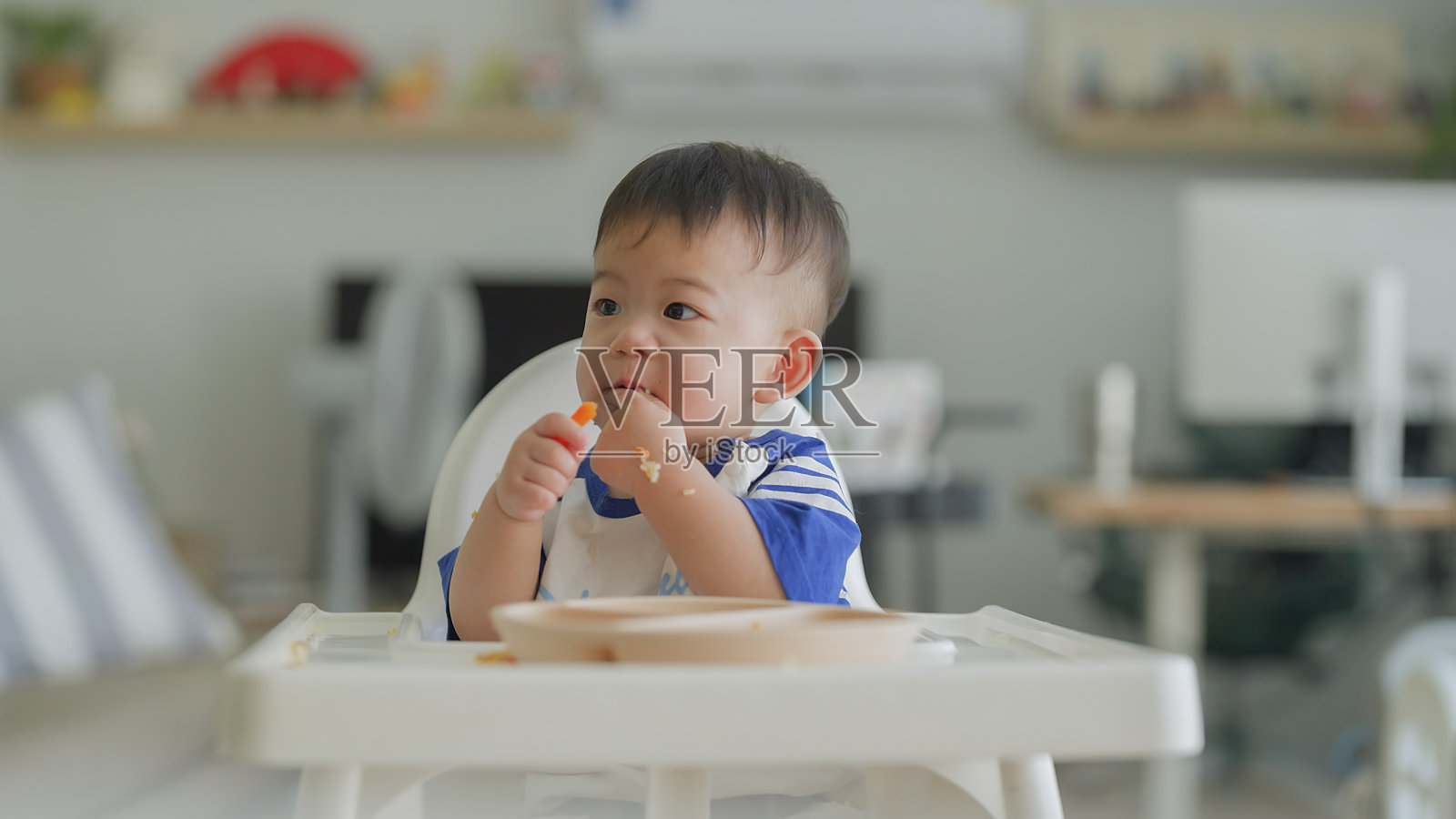 可爱的亚洲男婴坐在高脚椅上自己吃盘子里的食物，母亲的手在家里用勺子喂她的男婴，婴儿里程碑，成长和发展的概念照片摄影图片