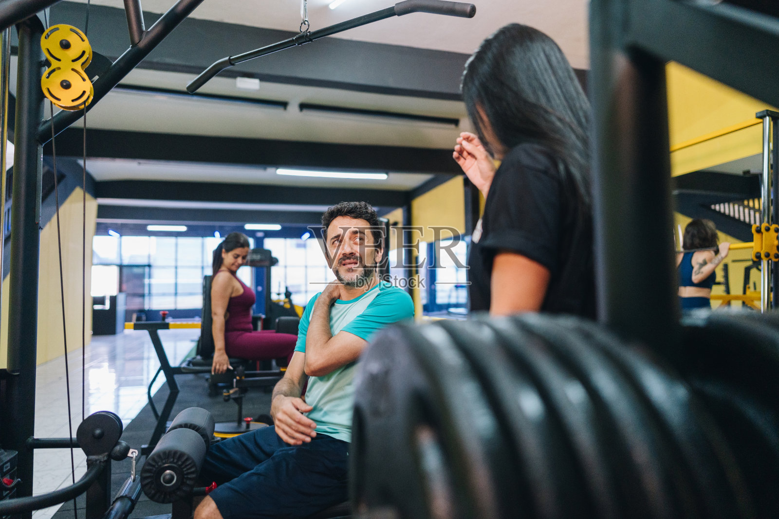 一个成熟的男人感到脖子疼，在健身房和健身教练聊天照片摄影图片