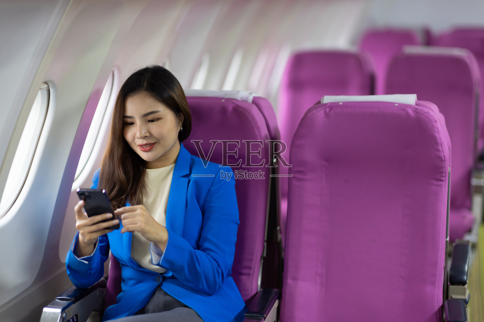 在飞机上玩手机的女商人。亚洲女游客在飞机上使用智能手机，女性在飞机上拿着移动设备玩游戏或工作。照片摄影图片