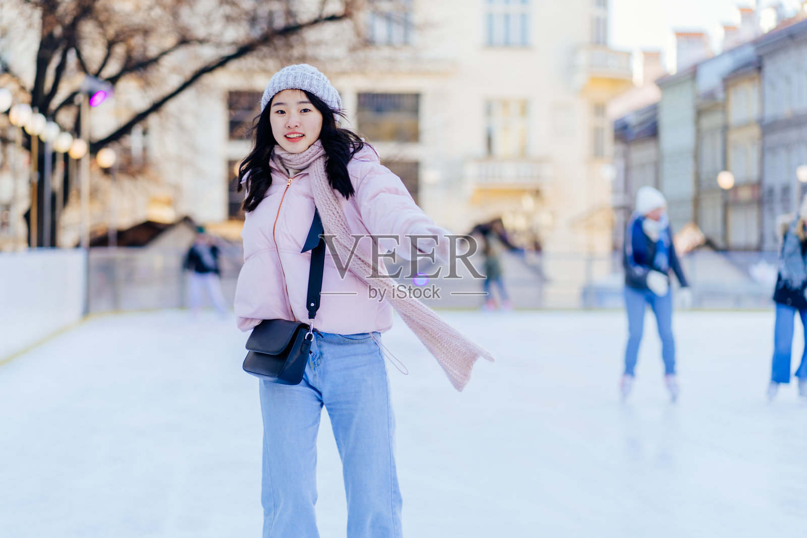 一位年轻的亚洲女性穿着粉粉色的夹克，戴着暖和的针织帽、围巾和手套，在冬天的户外溜冰场上滑冰。照片摄影图片