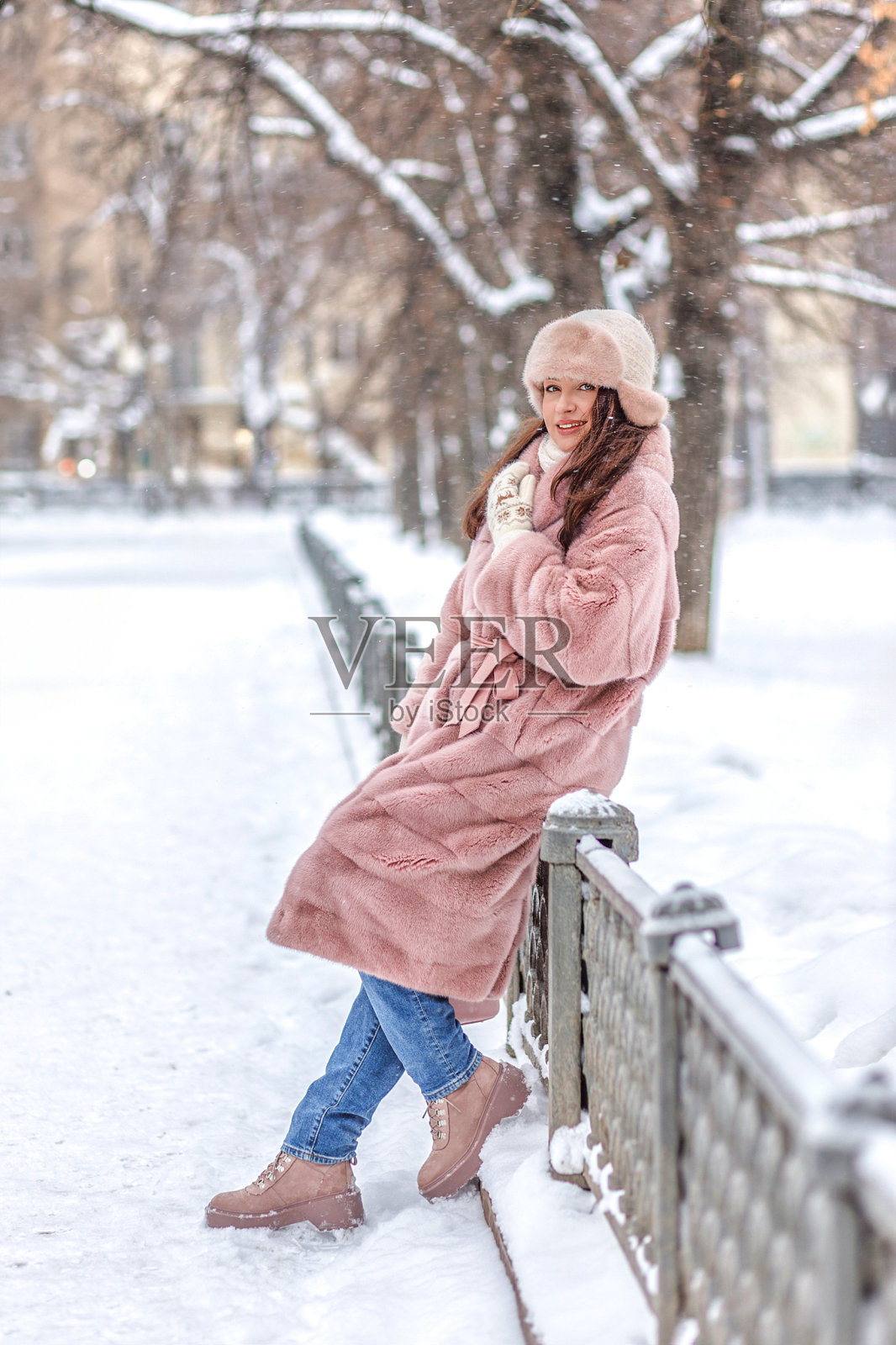 街上的肖像。优雅时髦的白人女子身穿粉色皮草大衣，头戴带耳盖的粉色蓬松帽子，走在新年的城市里，圣诞装饰照片摄影图片