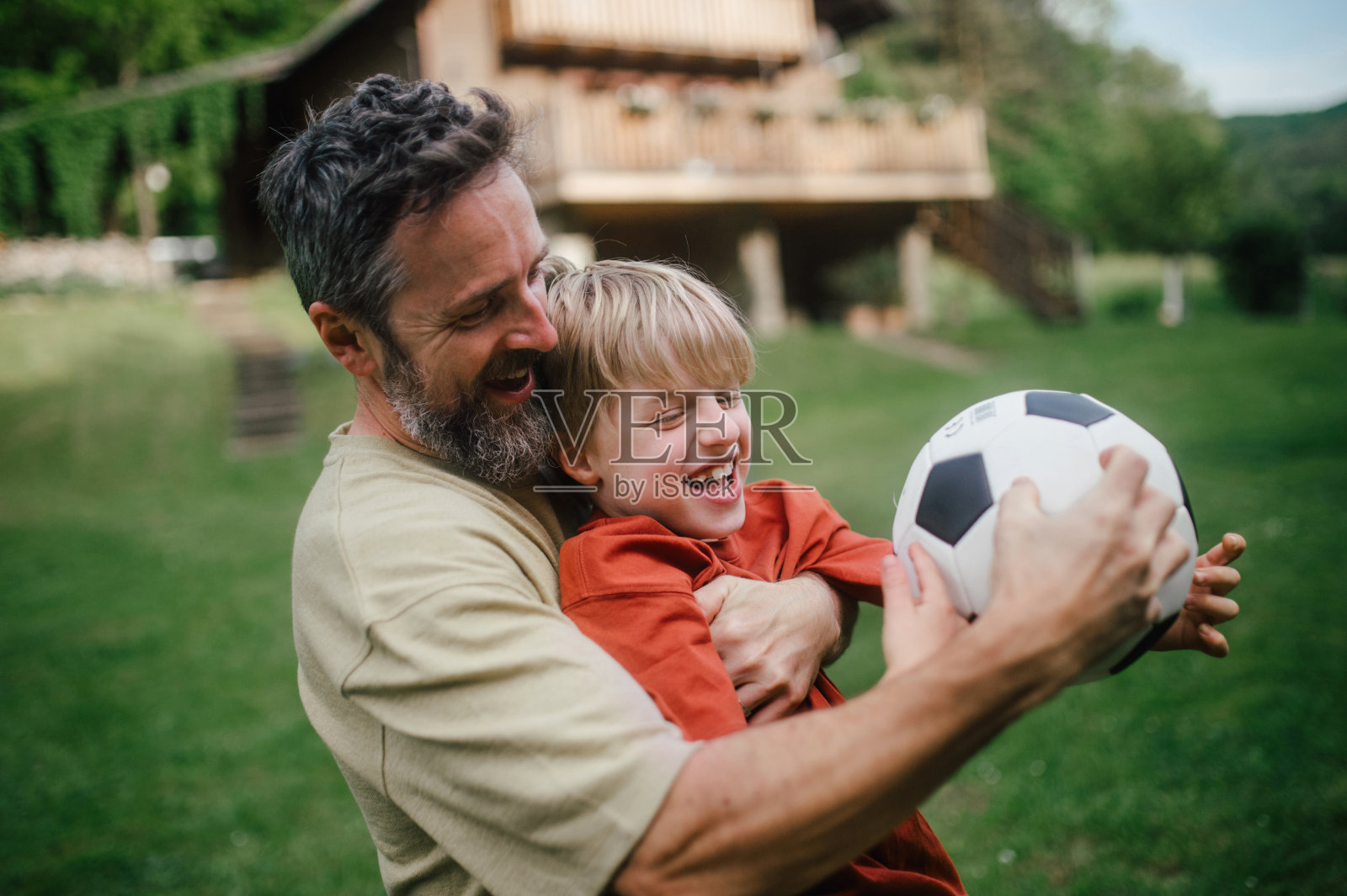爸爸和小儿子玩得很开心，踢足球，跑步，互相抓对方。在他们房前的草坪上玩耍。父亲和父亲节。照片摄影图片