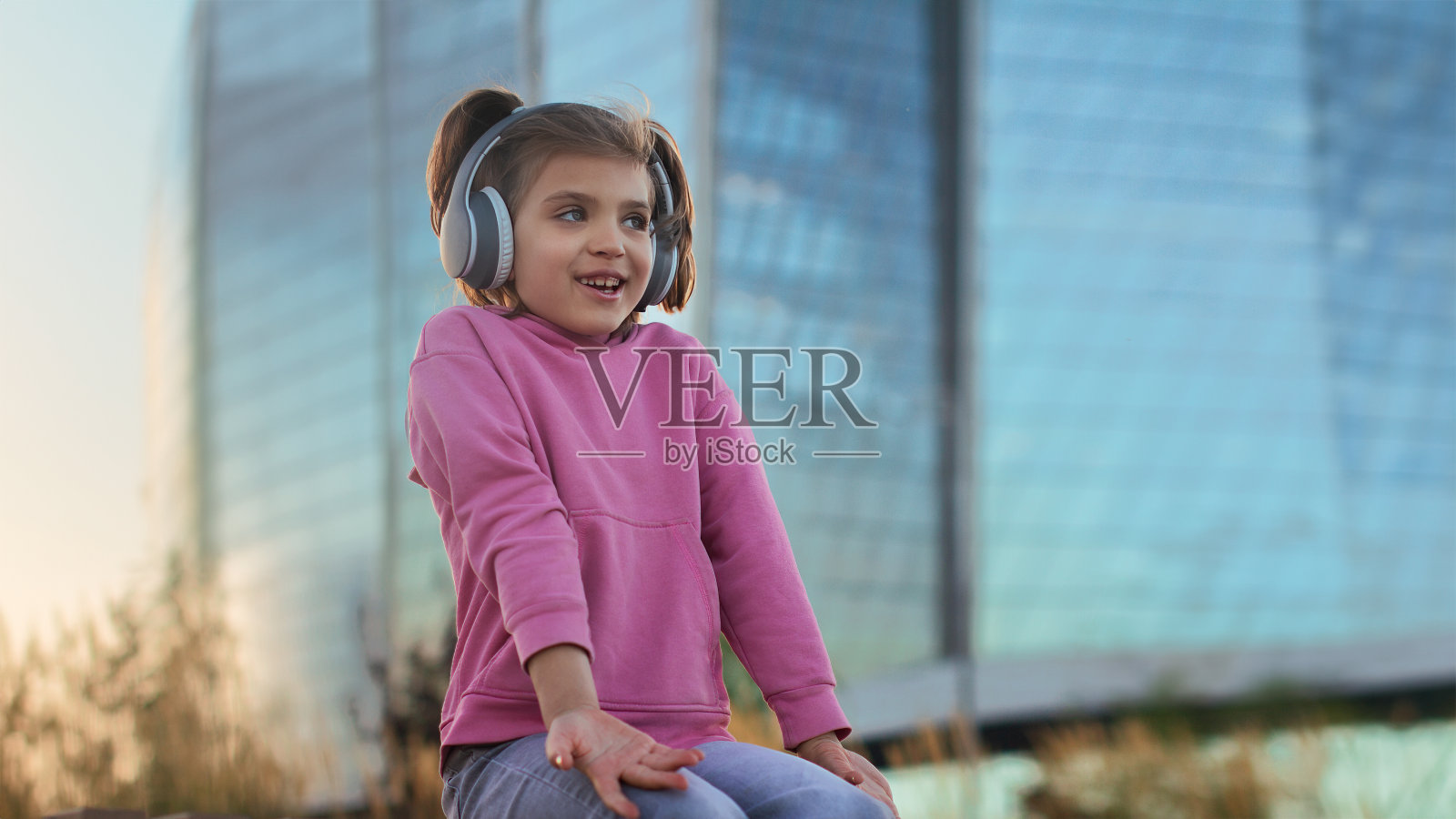 6-7岁穿着粉色连帽衫，戴着耳机在城市里听音乐的有趣女孩的肖像，生活方式照片摄影图片