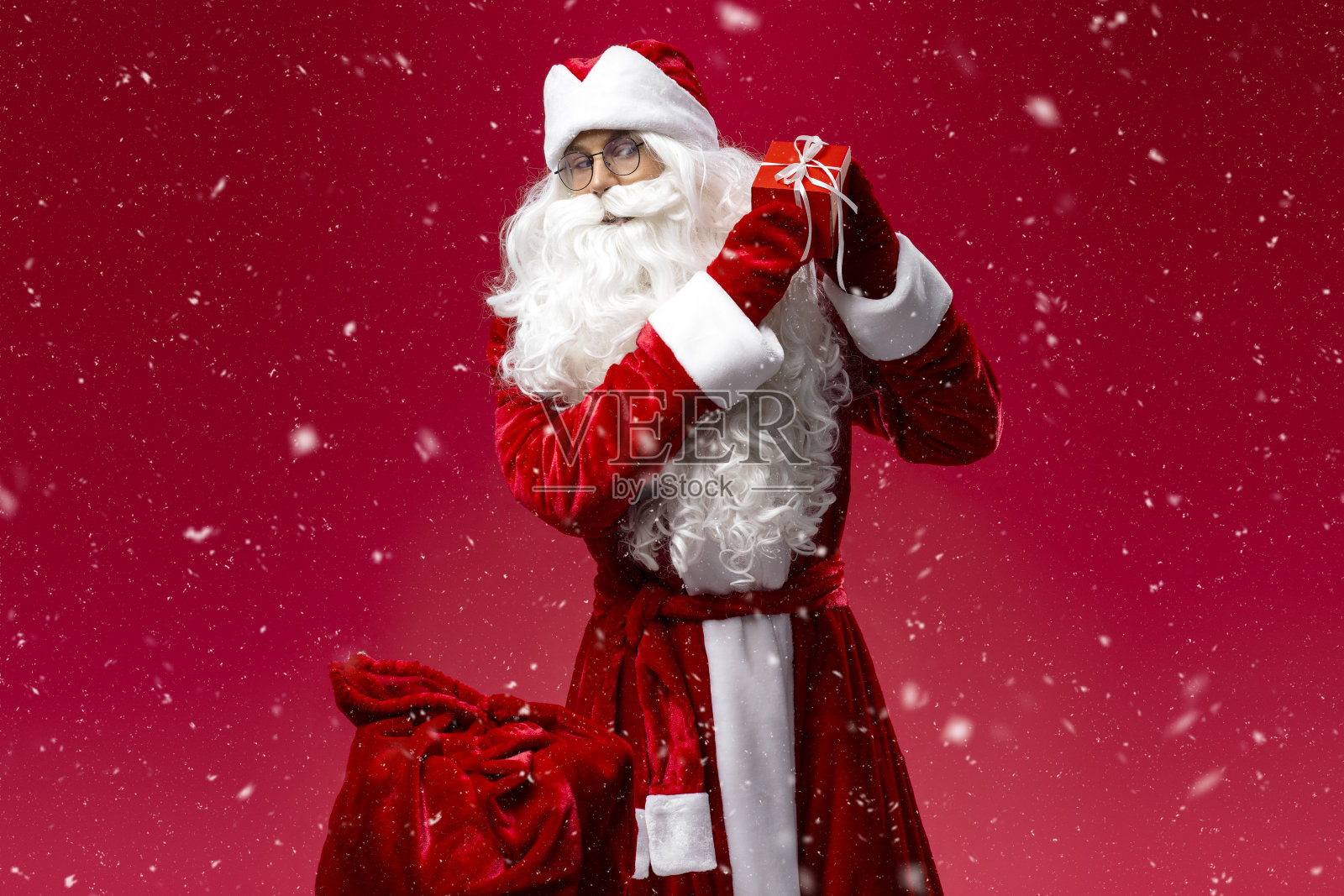 圣诞老人拿着礼盒看着相机孤立的红色背景与雪照片摄影图片