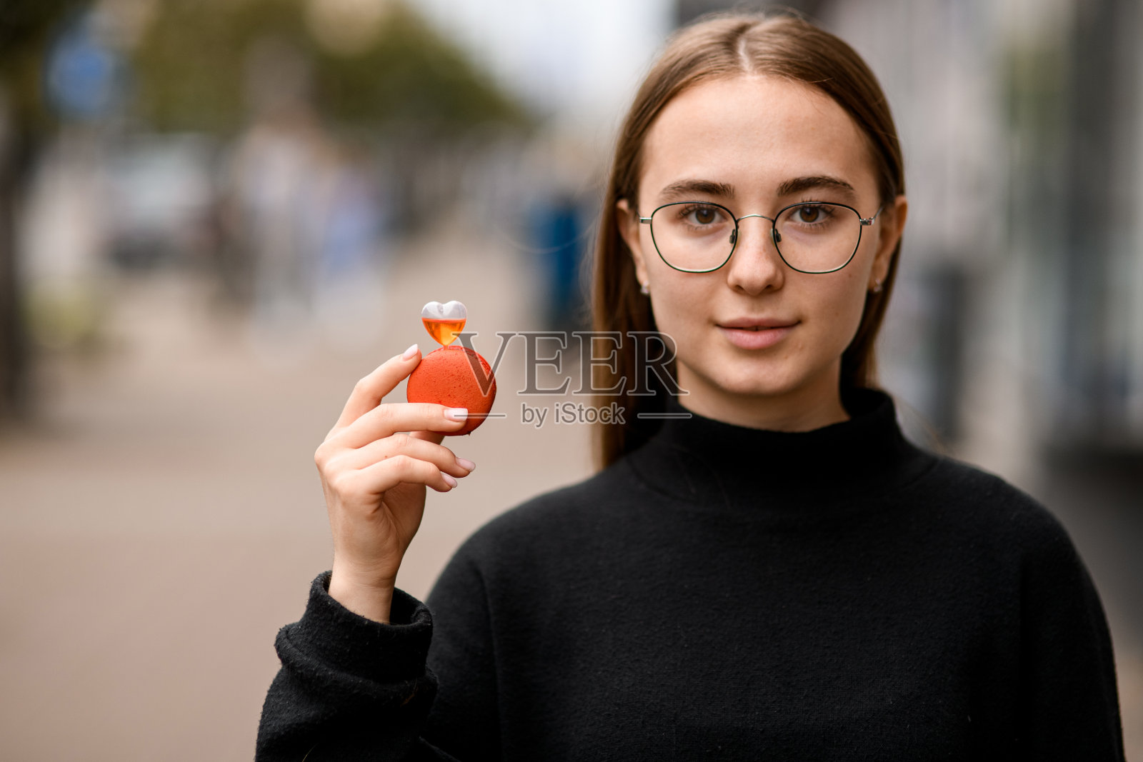 戴眼镜的漂亮的棕色眼睛的女孩手里拿着红色的法国马卡龙照片摄影图片