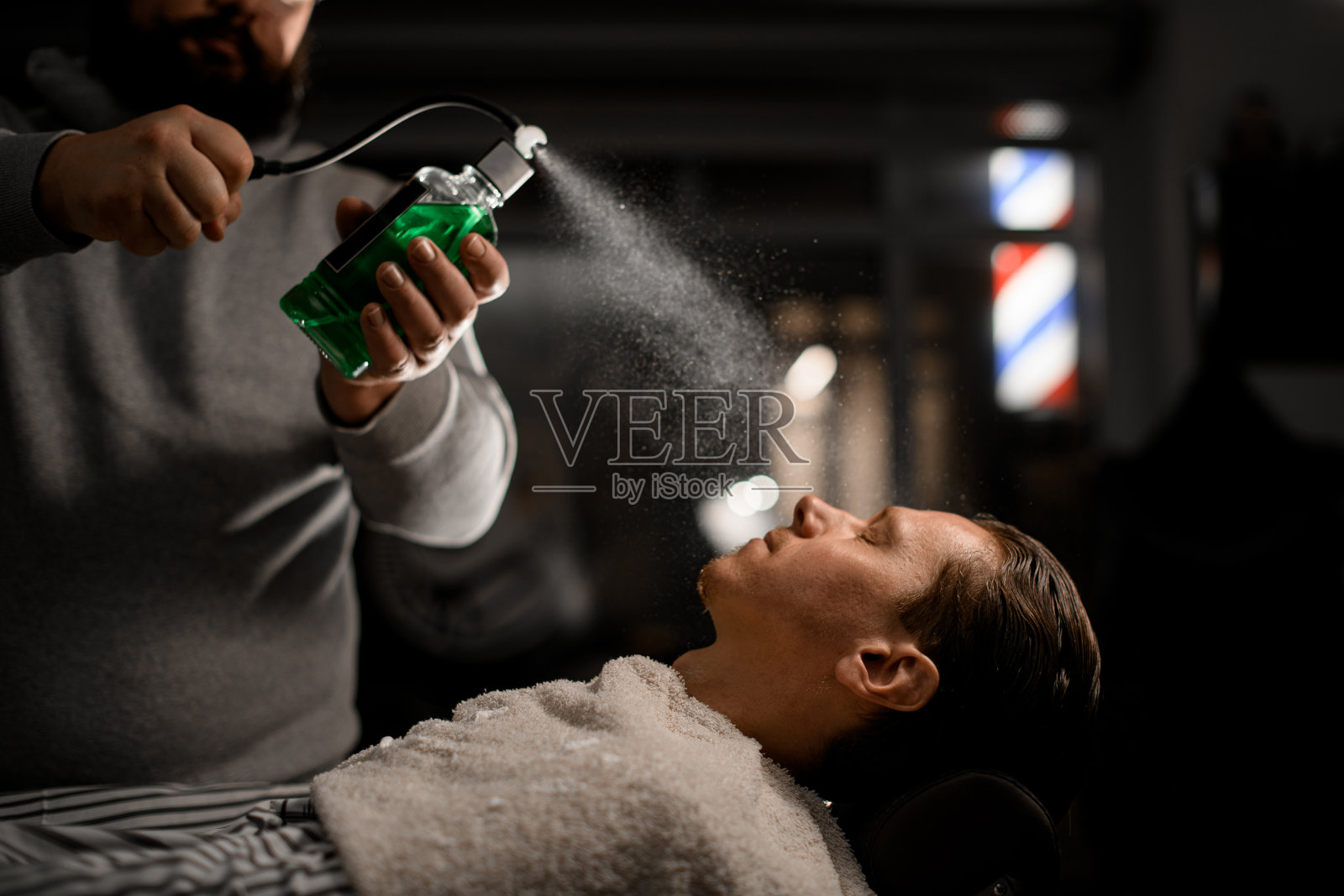 在老式理发店里，一位理发师从复古设计的时尚瓶子里向顾客脸上喷洒绿色古龙水照片摄影图片