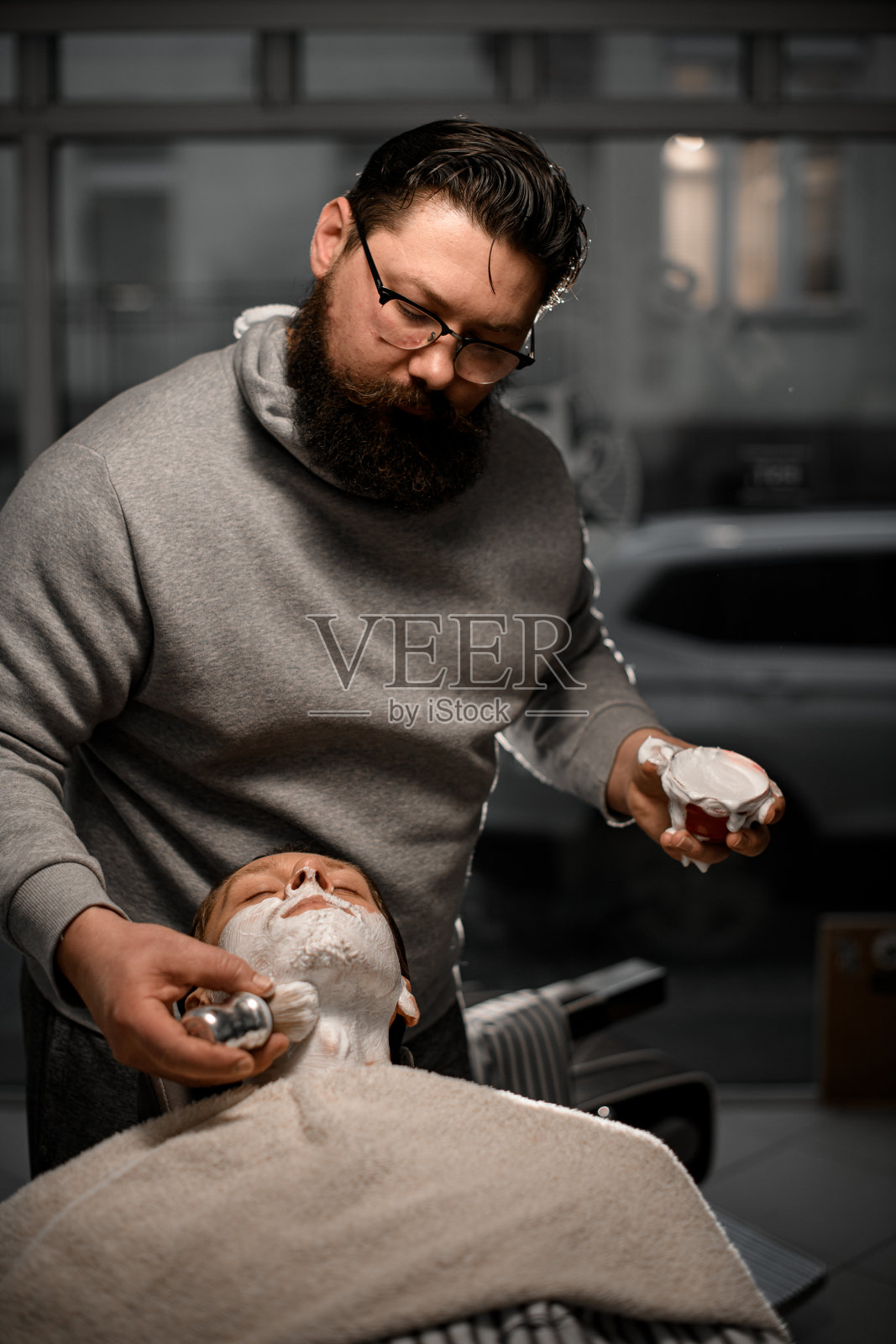 有胡子的时尚理发师在理发店用刷子将剃须泡沫涂在客户脸上照片摄影图片