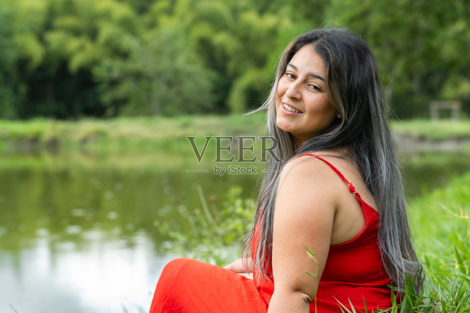 一个拉丁女人坐在湖边望着一边，脸上带着灿烂的笑容。红色的裙子照片摄影图片