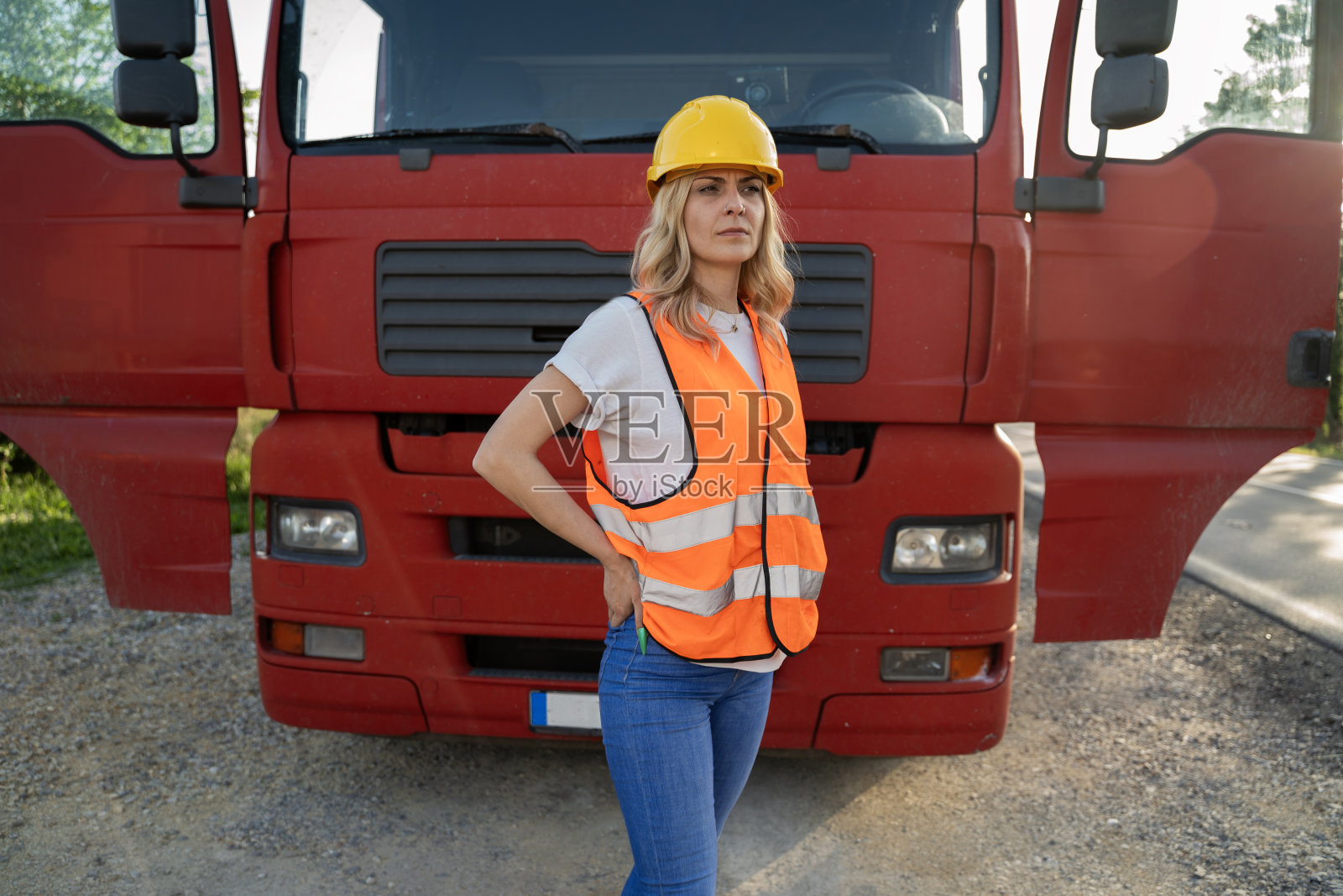 职业女卡车司机骄傲地站在红色卡车前。穿戴安全背心和安全帽照片摄影图片
