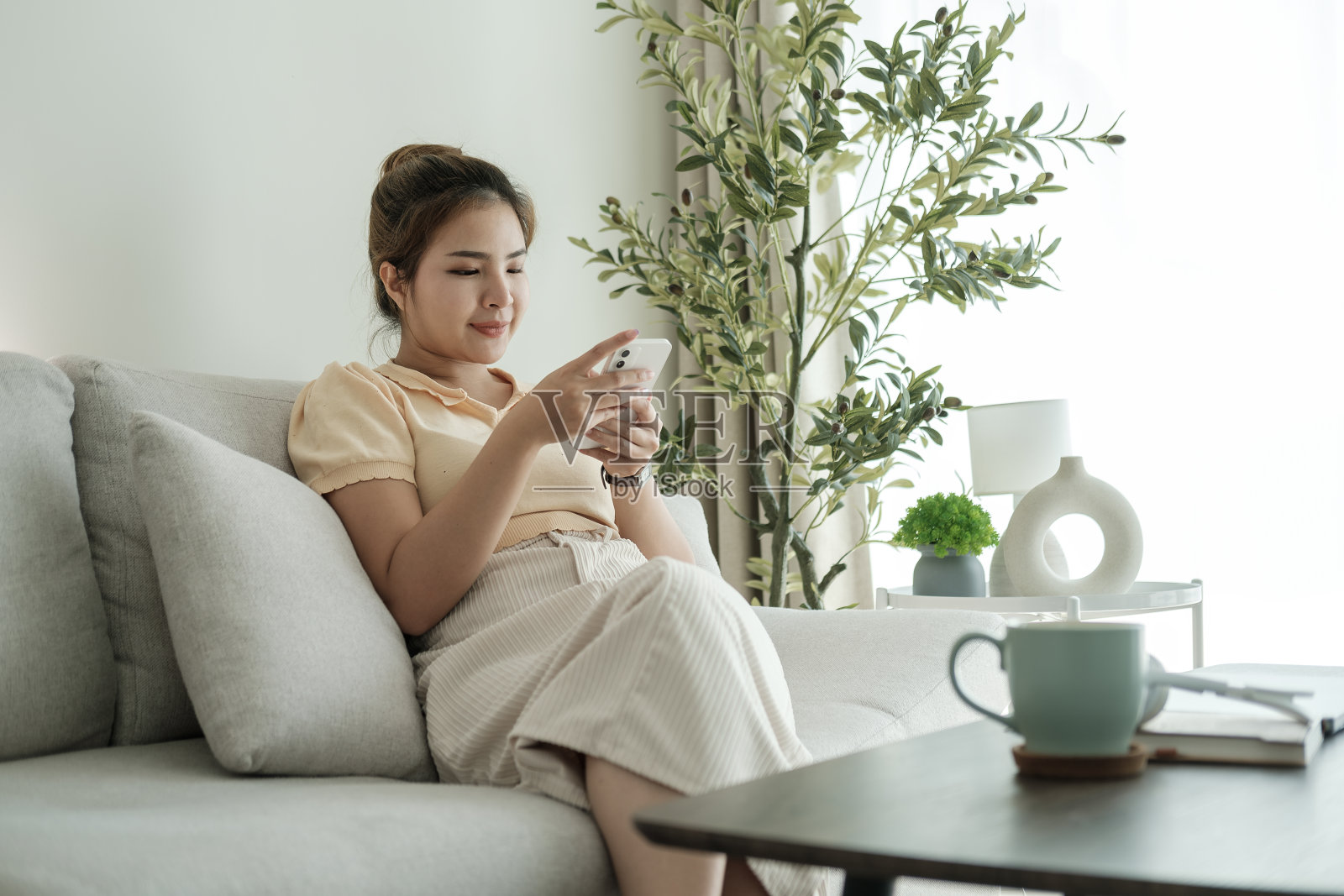 漂亮的年轻女子穿着休闲的衣服，坐在舒适的客厅的沙发上玩手机照片摄影图片
