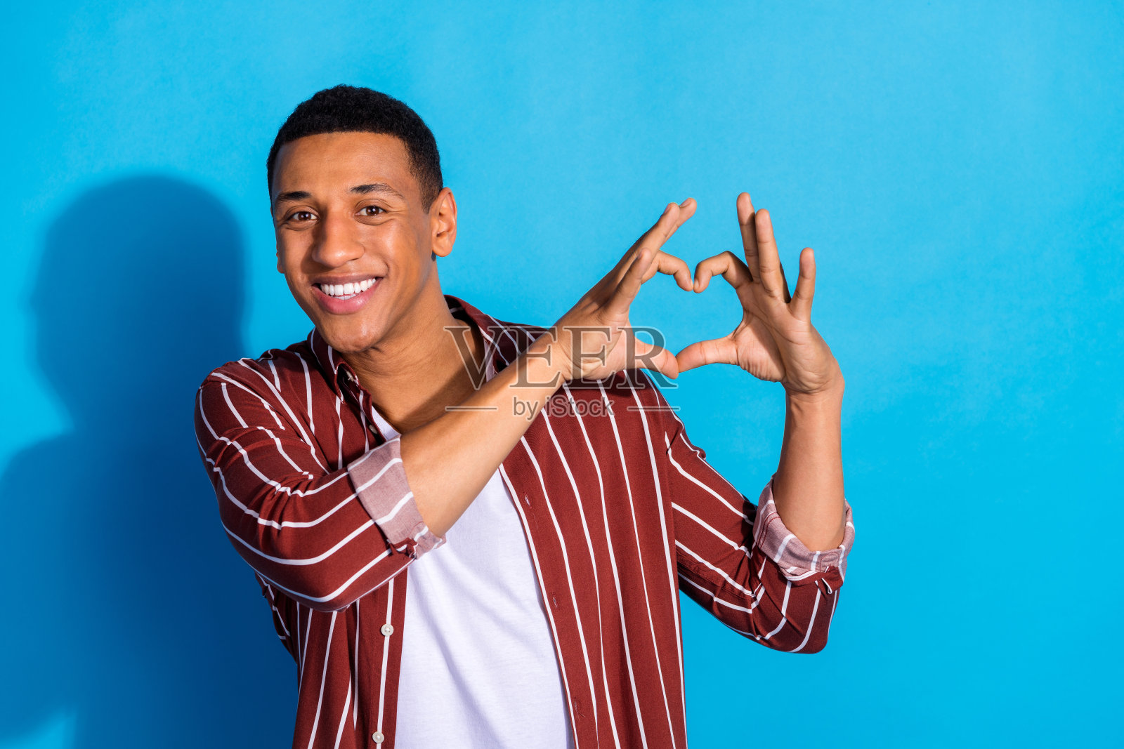 照片好心情帅哥穿条纹时尚衬衫在白色t恤手指显示心脏孤立在蓝色背景照片摄影图片