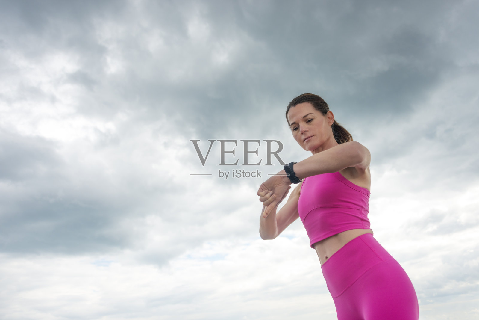 一个运动的女人在看她的健身追踪表，她穿着粉红色的运动服，衬着戏剧性的天空。照片摄影图片