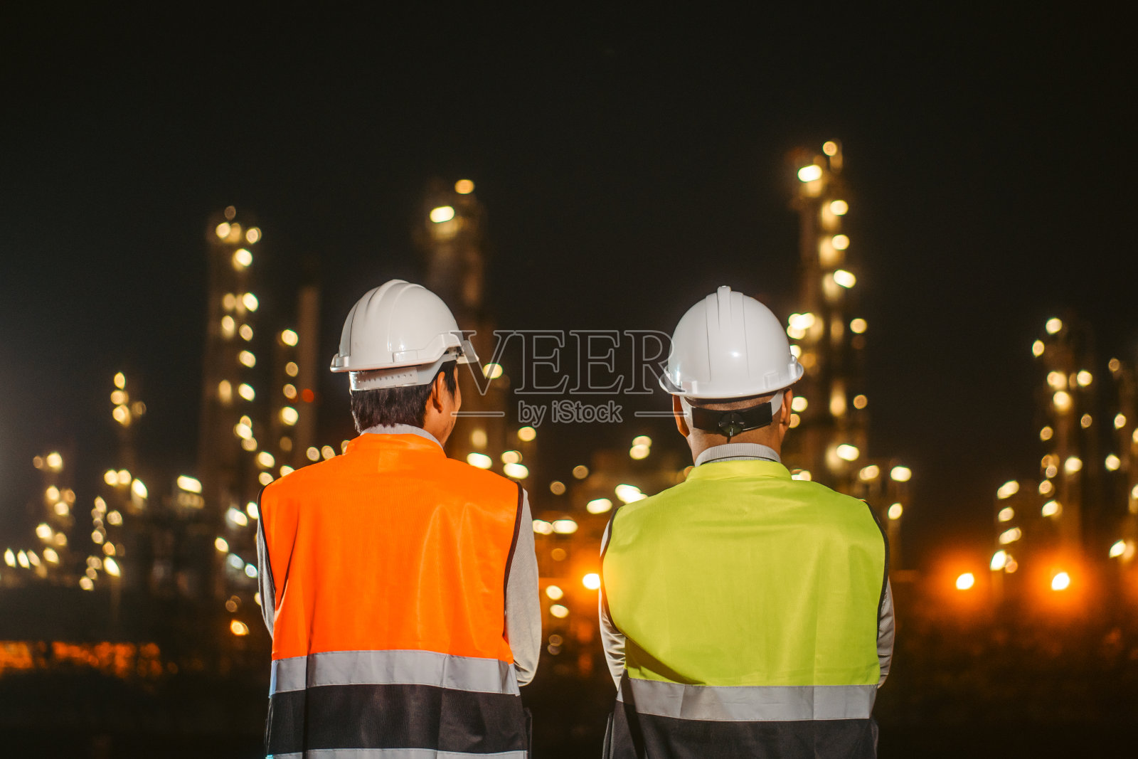 两个戴着安全帽的亚洲工程师站在炼油厂前。工业园区天然气石化。工厂储油罐和管道。工人们在炼油厂建筑大楼里工作照片摄影图片