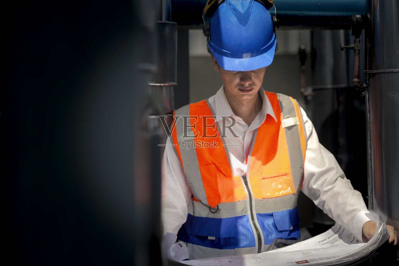 一名工业工人戴着高能见度装备和蓝色头盔，一丝不苟地检查着错综复杂的机械网络。工程师致力于维护操作效率和安全。照片摄影图片