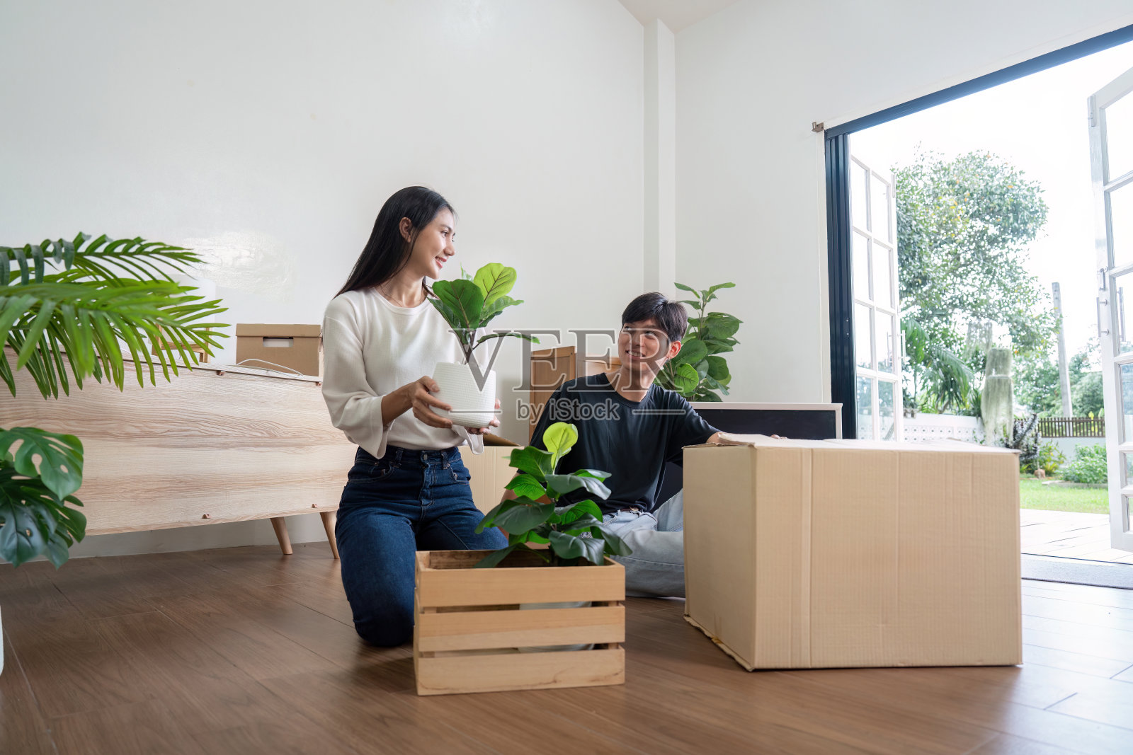 幸福的夫妇搬进新家，打开箱子，在明亮的现代客厅里安排植物，大窗户和木地板照片摄影图片